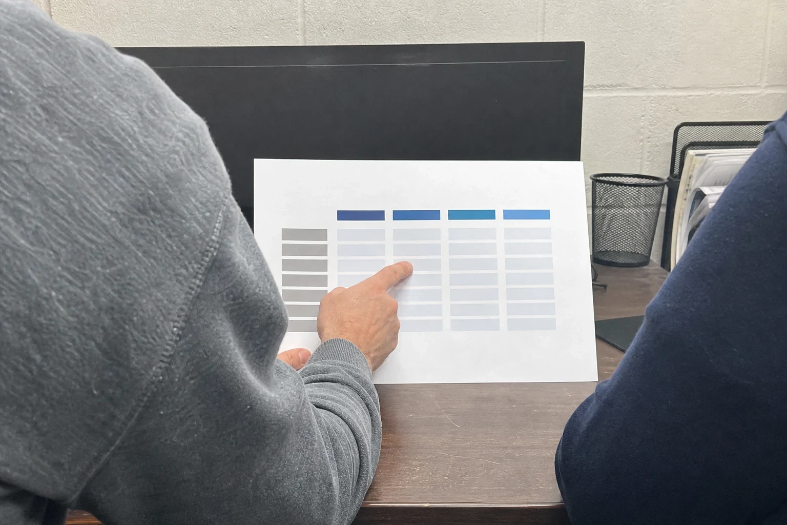 Over-the-shoulder view of a person in a gray sweatshirt pointing at a printed chart on a wooden desk in a small cinderblock back office