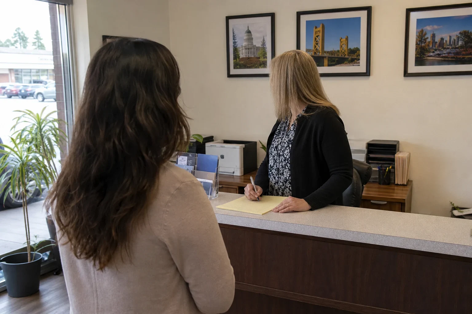 Client with long brown hair stands at an agency reception counter while an agent writes on a notepad, with framed Sacramento photos on the wall and plants by the window